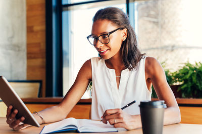 Young woman using digital tablet while writing on book