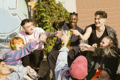 Happy non-binary friends celebrating with champagne while sitting together in city at sunny day