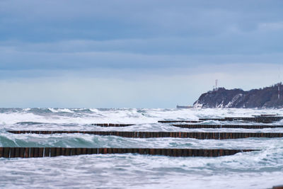 Scenic view of blue sea with foaming waves, cloudy sky and vintage long wooden breakwaters in sea