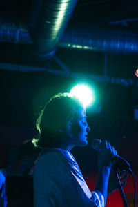 Confident lady with guitar singing in mic while performing song in bright club with neon light