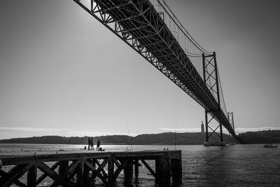 Distant view of men on pier at tagus river below april 25th bridge