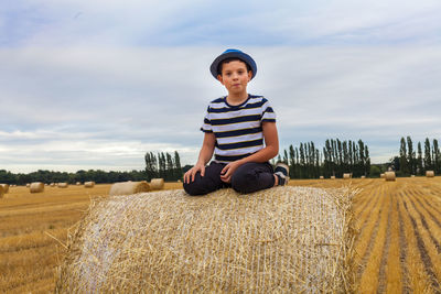 Portrait of cute boy sitting on hay bale against cloudy sky