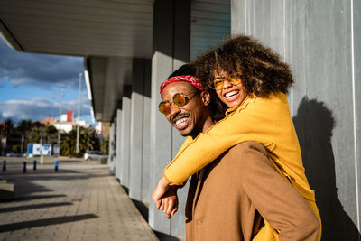 Cool african american man in sunglasses and coat giving piggyback ride to bright laughing girlfriend with afro curly hair looking at camera while spending time on street