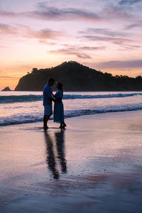 Rear view of man standing at beach against sky during sunset