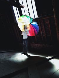 Woman holding colorful umbrella while standing by window in darkroom