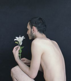Side view of shirtless young man holding flower while crouching against wall