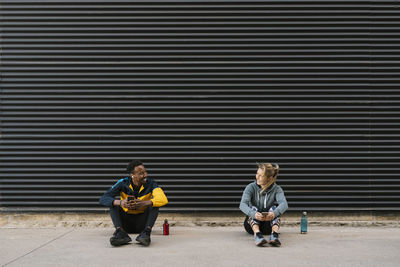 Smiling male and female athlete with mobile phone looking at each other while sitting against wall