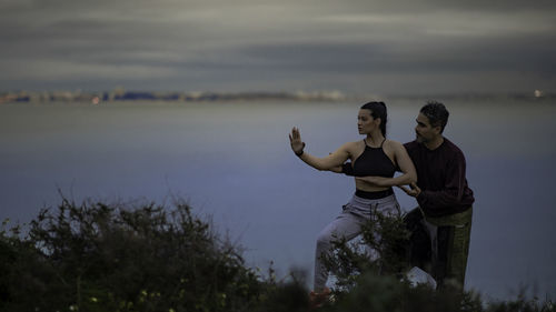 Man instructing woman while exercising against lake