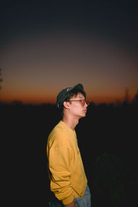 Side view of young man wearing sunglasses standing against sky at dusk