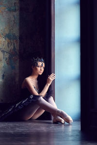 Full length of thoughtful young ballet dancer sitting at doorway against wall