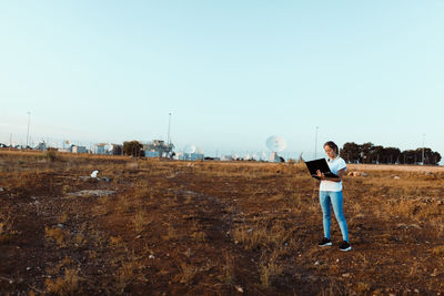 Full length of woman holding laptop standing on field against clear sky