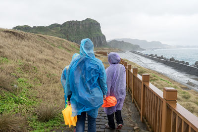 Rear view of people standing by railing and sea against sky during rainy day