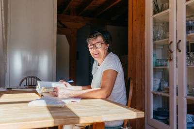 Focused middle aged female in glasses with pen in hand sitting at table with heap of books and papers and studying professional materials at home