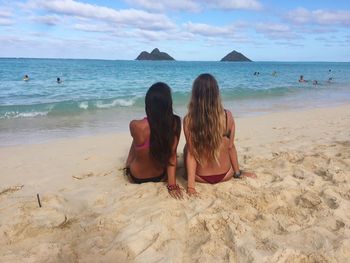 Rear view of female friends sitting on sand at beach against sky