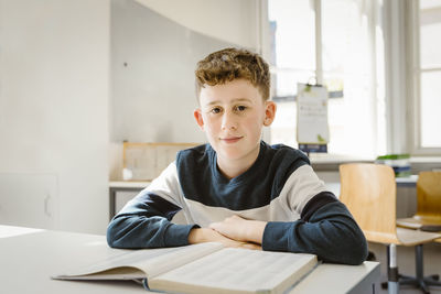 Portrait of schoolboy sitting with book at desk in classroom