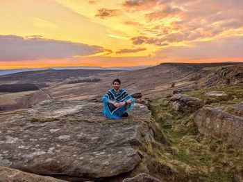 Rear view of man sitting on rock against sky during sunset