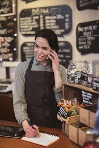 Smiling female owner using phone while writing in note pad at store counter