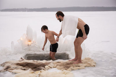 Full length of man holding hand of son taking ice bath at frozen lake