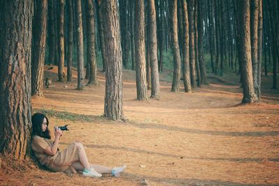 Woman photographing through camera while sitting on field at forest