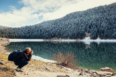 Side view of mid adult man photographing while crouching by lake against sky