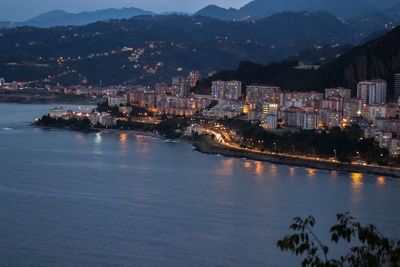 Illuminated buildings by sea against sky at dusk