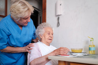 Caring nurse in uniform helping aged female patient sitting at table with bowl of soup in light kitchen at home