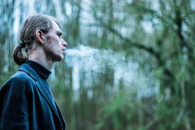 Side view of young man smoking while standing against trees in forest