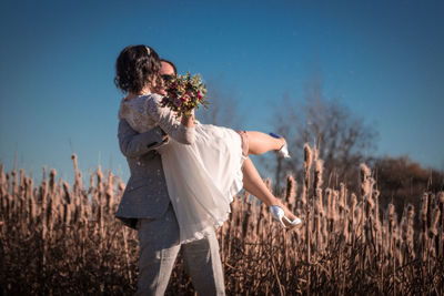 Groom carrying bride holding bouquet on land against sky