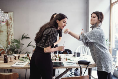 Young woman smelling perfume fragrance from female colleague's wrist while standing at workshop