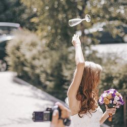 Cropped hand of man filming bride throwing champagne flute against plants
