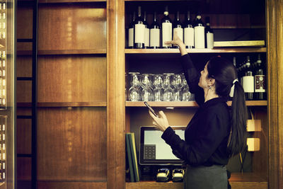 Side view of owner checking list of wine bottles while standing in cafe