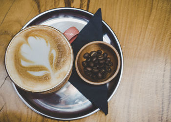 Fresh cappuccino on metal plate on wooden surface. decorated with coffee beans in small wooden bowl.