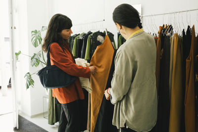 Female sales staff assisting woman while choosing dress from rack at clothes store