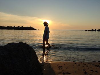 Girl wading in sea against sky during sunset