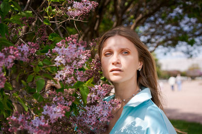 A woman stands in front bush with purple flowers. she is wearing blue shirt and has her hair down