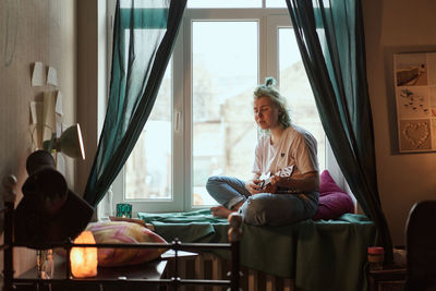 Woman playing guitar while sitting on window sill at home