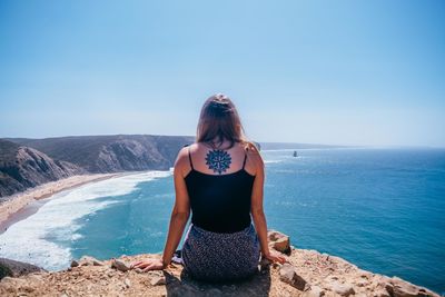 Rear view of young woman sitting on cliff against sea