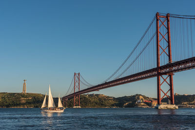 Sailboat sailing in tagus river by 25 de abril bridge against clear blue sky