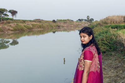 Portrait of young woman standing by lake against sky