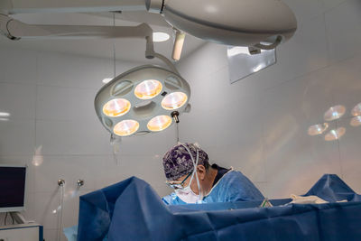 Professional senior male surgeon in mask and uniform doing operation under lamp in operating room