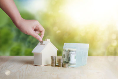 Cropped hand putting coin in piggy bank by currency on table against wall