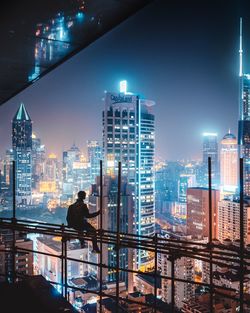 Rear view of silhouette man sitting against illuminated buildings on construction site in city at night