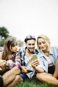 Young woman using mobile phone while sitting on grass against sky