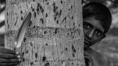 Portrait of shirtless boy holding feather while standing behind tree trunk