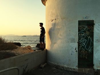 Side view of young man standing on wall by lighthouse against sea