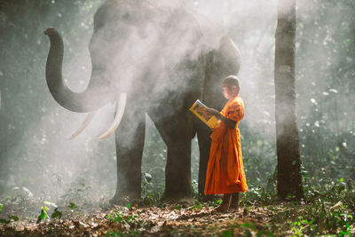 View of monk holding book standing by elephant in forest