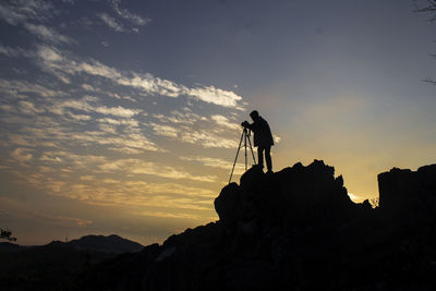 Silhouette man photographing on rock against sky during sunset