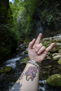 Close-up of tattooed hand gesturing against stream flowing in forest