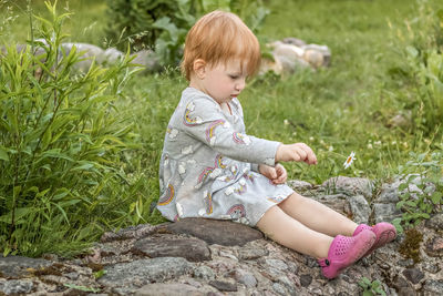 Little girl with red hair sits with a camomile in her hand in the garden among the greenery.