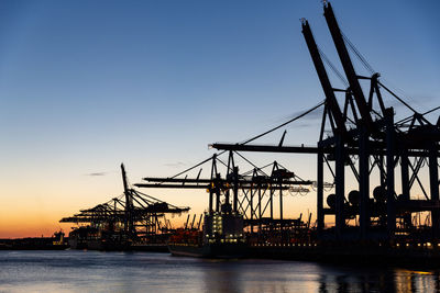 Silhouette cranes at harbor against sky during sunset